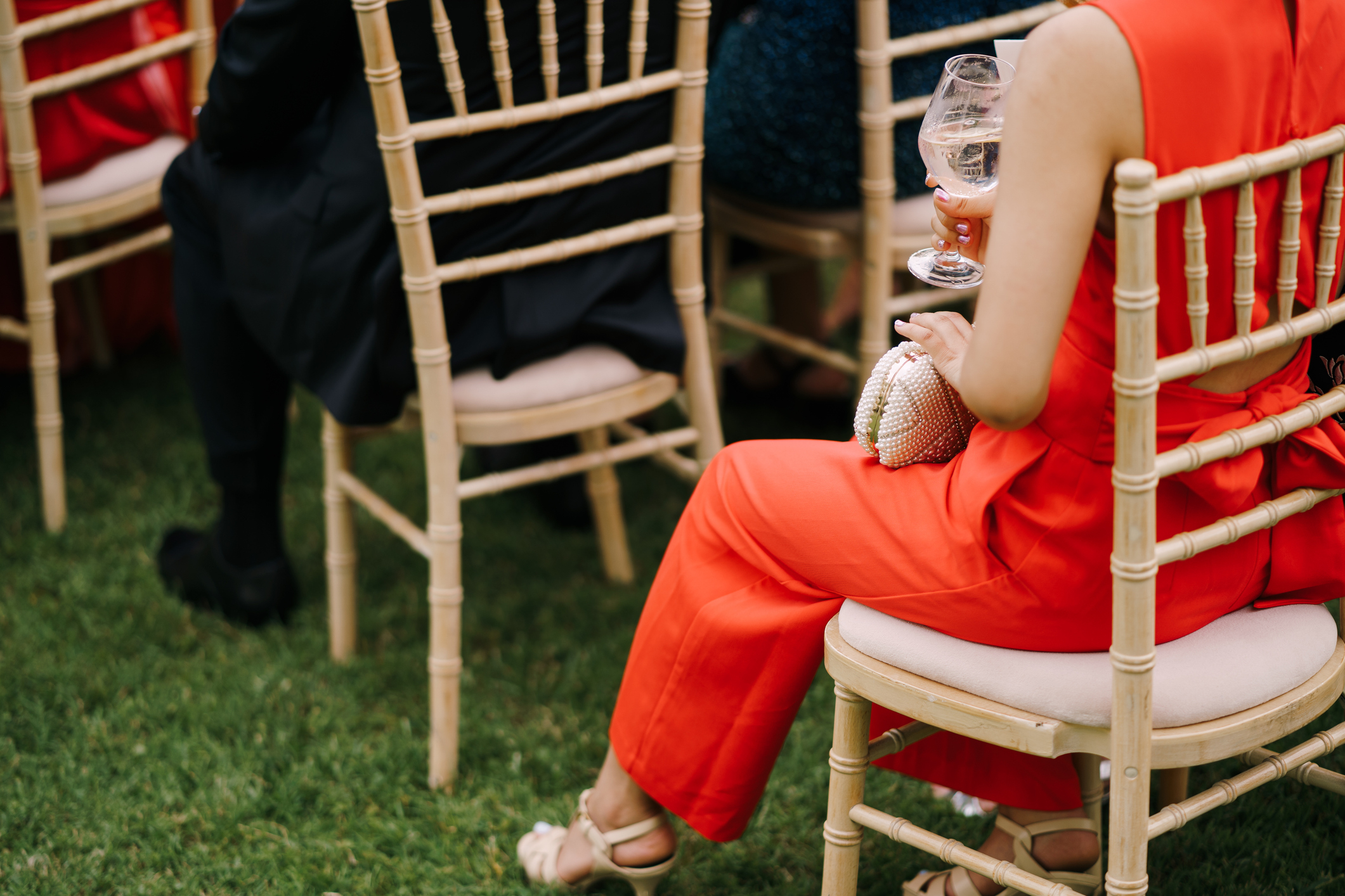 Woman in an elegant red jumpsuit sits on a wooden chair at an outdoor wedding, holding a drink and a small clutch bag