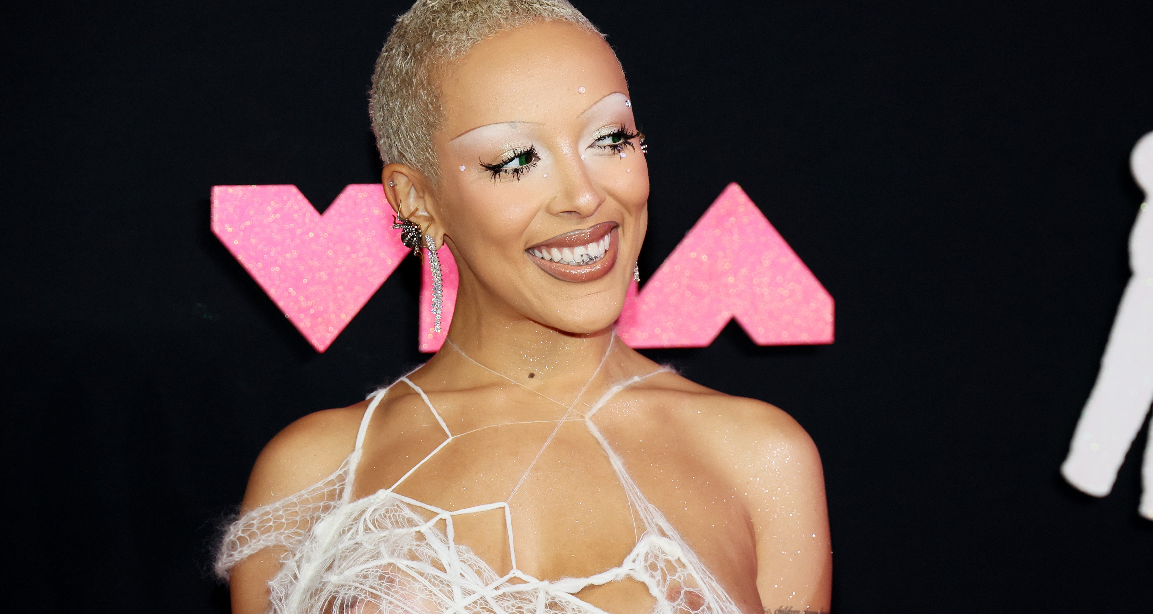 Doja Cat at the VMAs in a unique outfit with spider web-style strands covering her top. She has shaved hair and bold, creative makeup