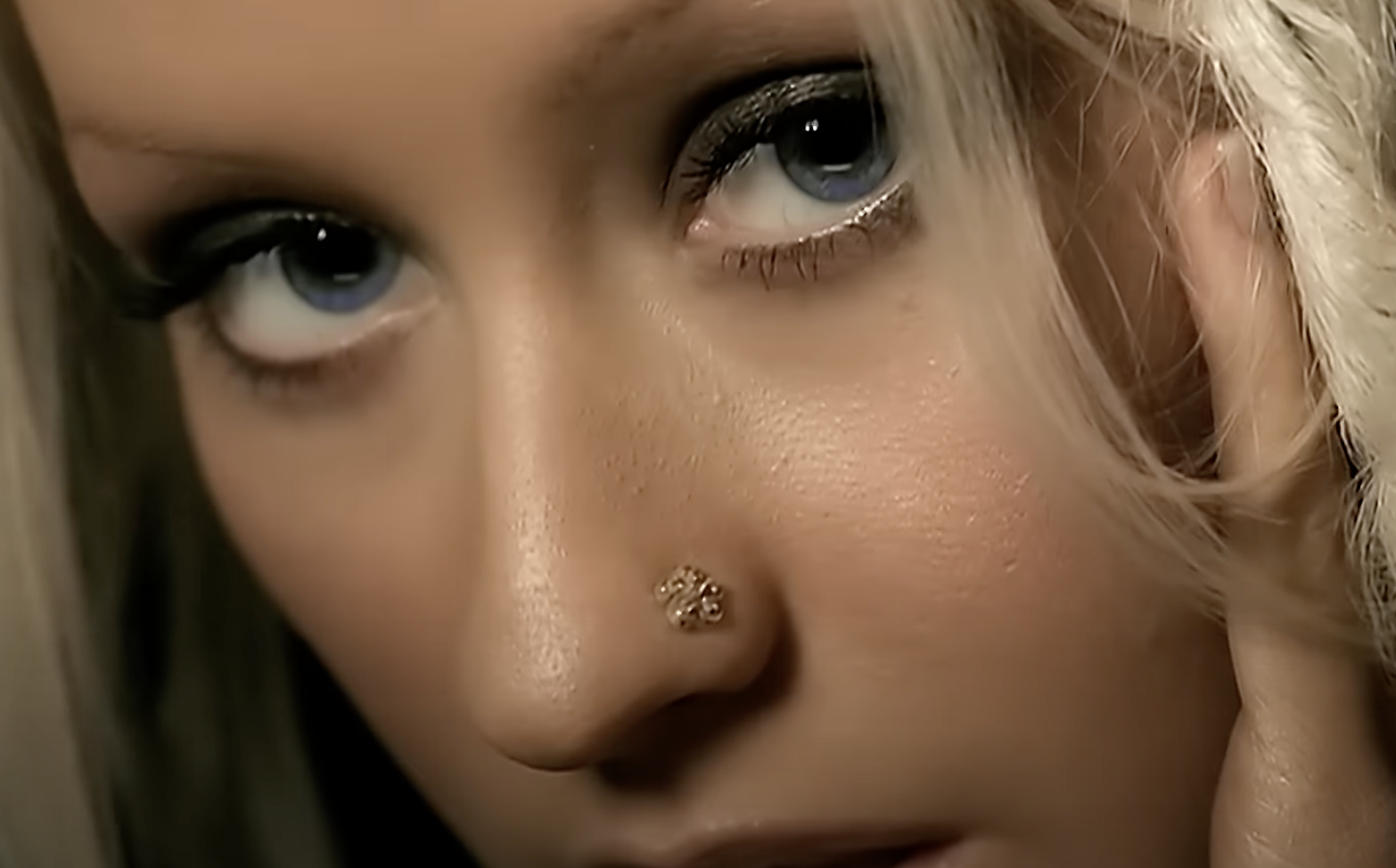 Close-up of Christina Aguilera's face with a jeweled nose ring and dramatic eye makeup, looking directly at the camera