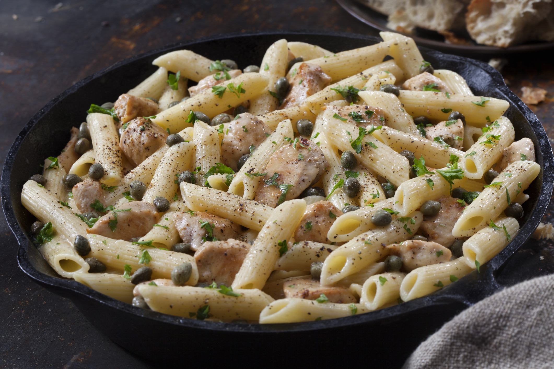 A skillet filled with penne pasta, chicken pieces, capers, and garnished with green herbs, placed on a dark table with bread in the background