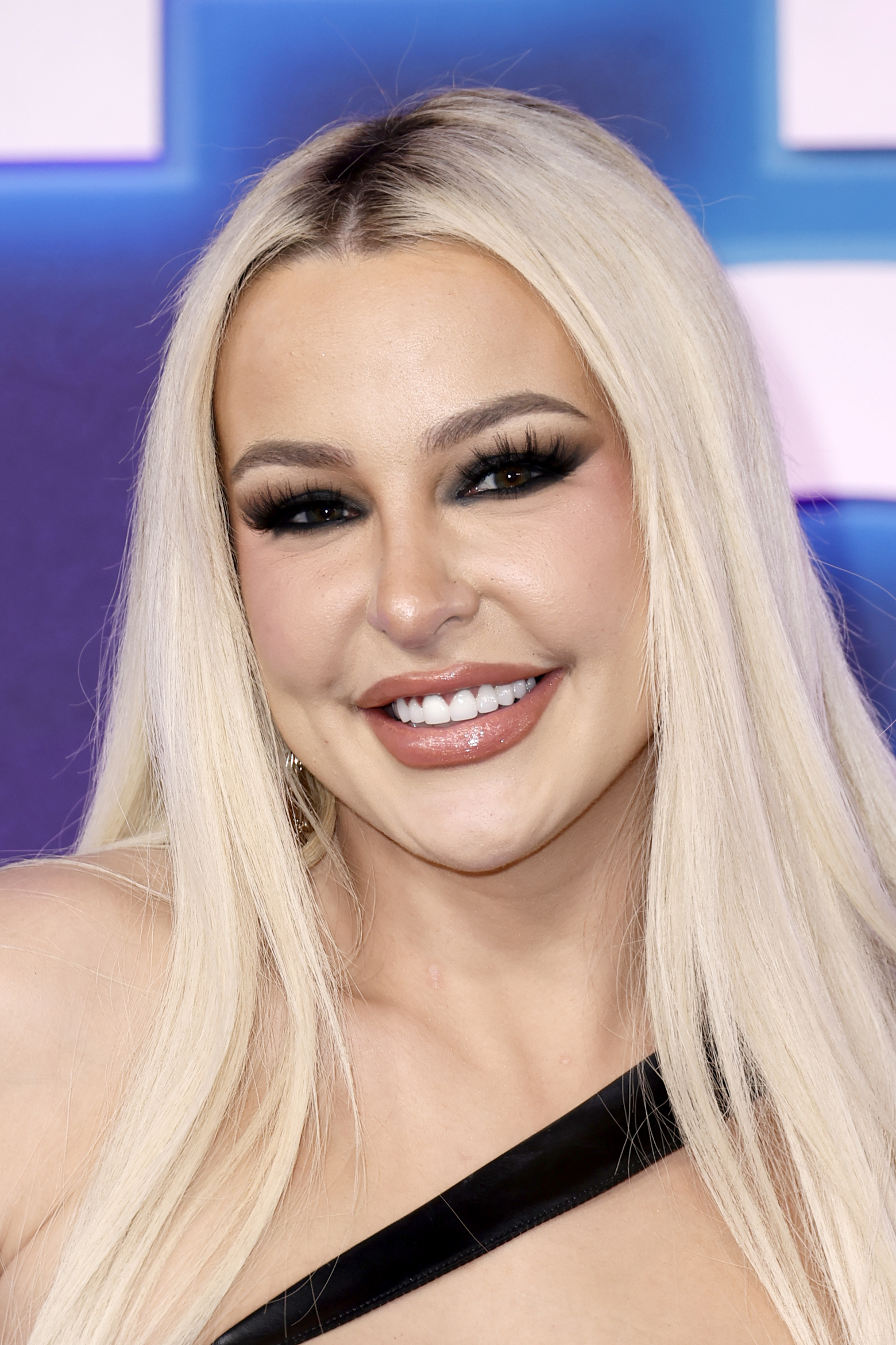 Tana Mongeau smiles at the camera, wearing a one-shoulder dress and dramatic eye makeup