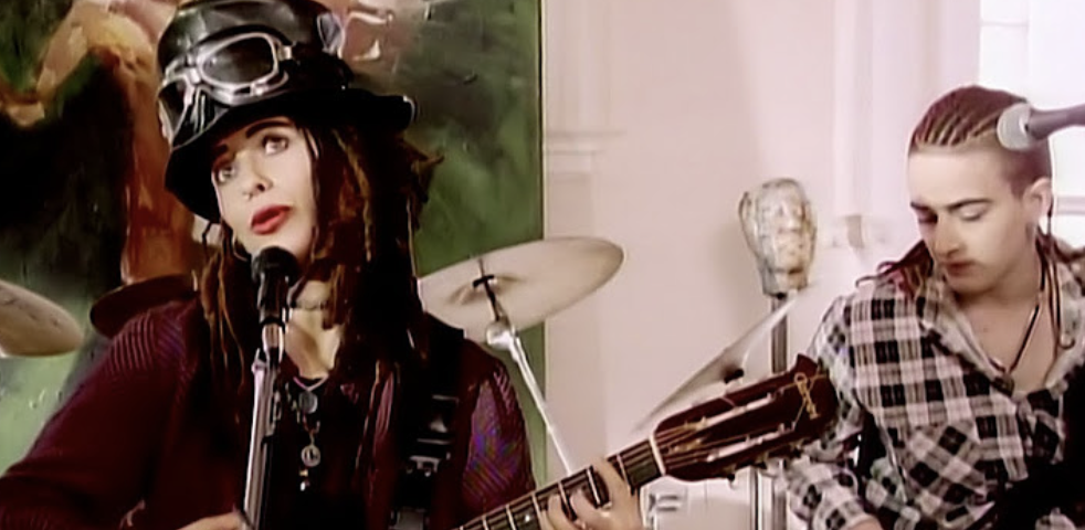 Boy George performs with drummer Jon Moss. Boy George wears a hat with goggles and has dreadlocks. Jon Moss has braided hair and wears a plaid shirt