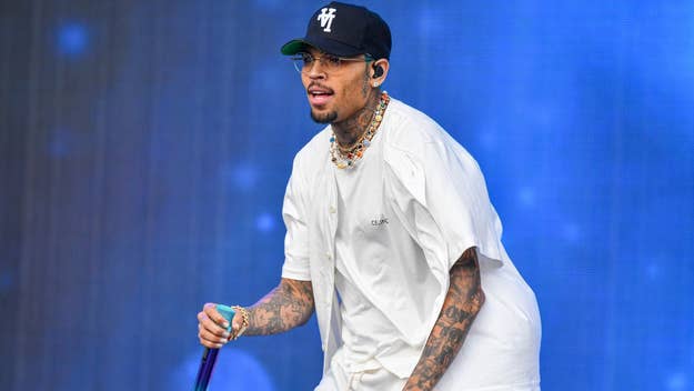 Chris Brown performing on stage, wearing a white button-up shirt, white pants, a black cap, and glasses
