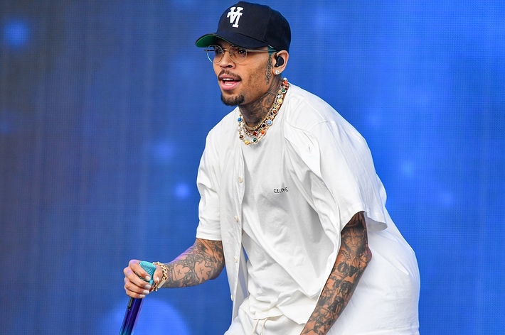 Chris Brown performing on stage, wearing a white short-sleeve shirt, hat, and glasses, with tattoos visible on his arms