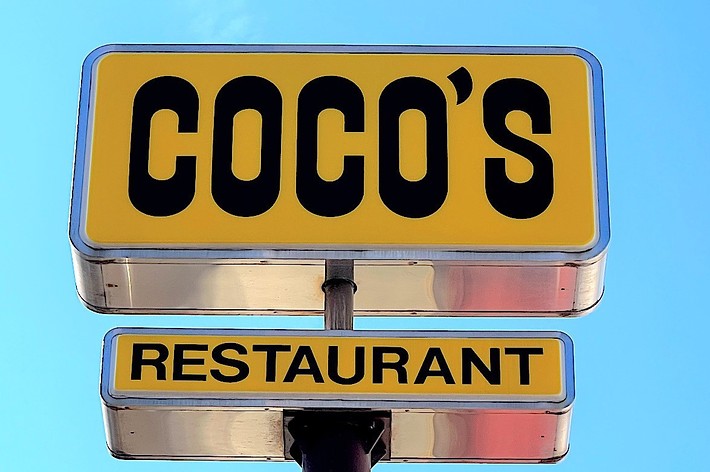 A sign for Coco’s Restaurant with bold black lettering on a bright background against a clear sky