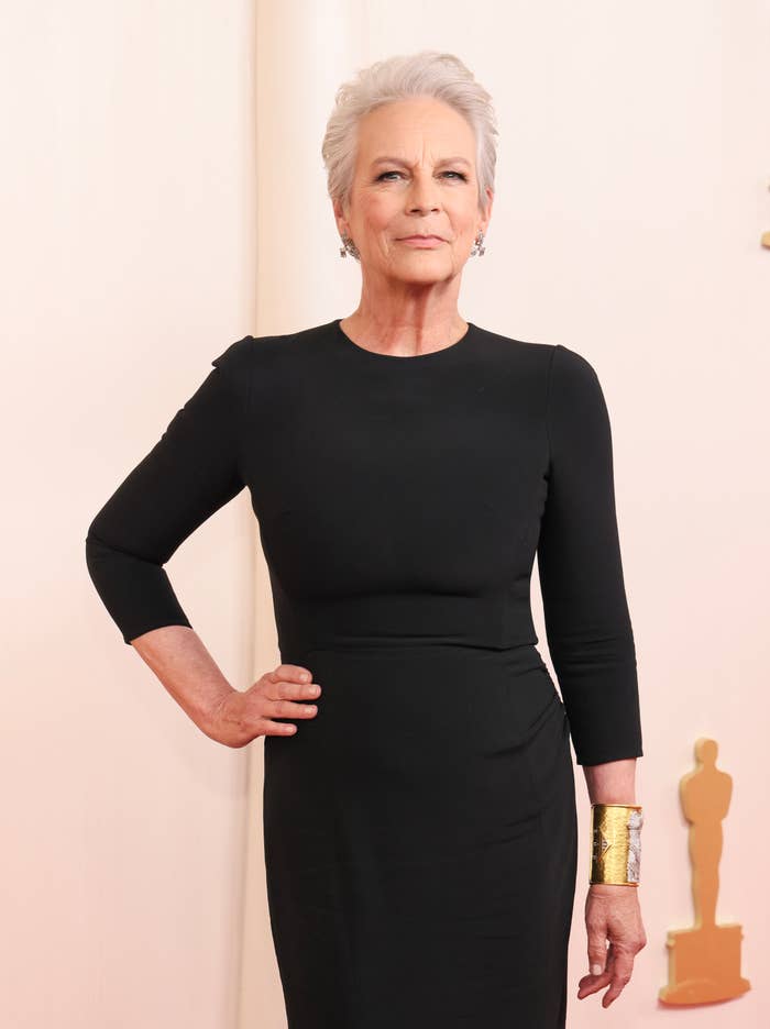 Jamie Lee Curtis poses on the red carpet in a fitted, long-sleeve black dress, accessorized with a wide gold cuff bracelet
