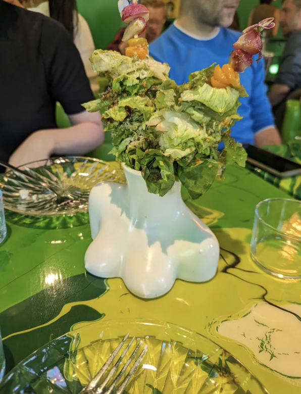 Salad served in a quirky white foot-shaped vase with colorful toppings, placed on a green table. Diners’ arms and a fork are visible in the background