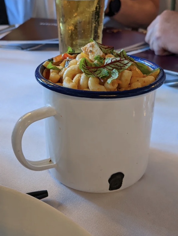 A tin cup filled with macaroni pasta garnished with herbs, positioned on a table with menu folders and a glass in the background