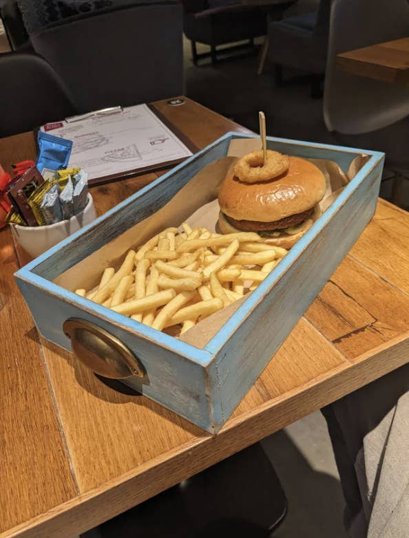 A burger garnished with an onion ring and a serving of fries are presented in a rustic blue wooden drawer on a table in a casual dining setting