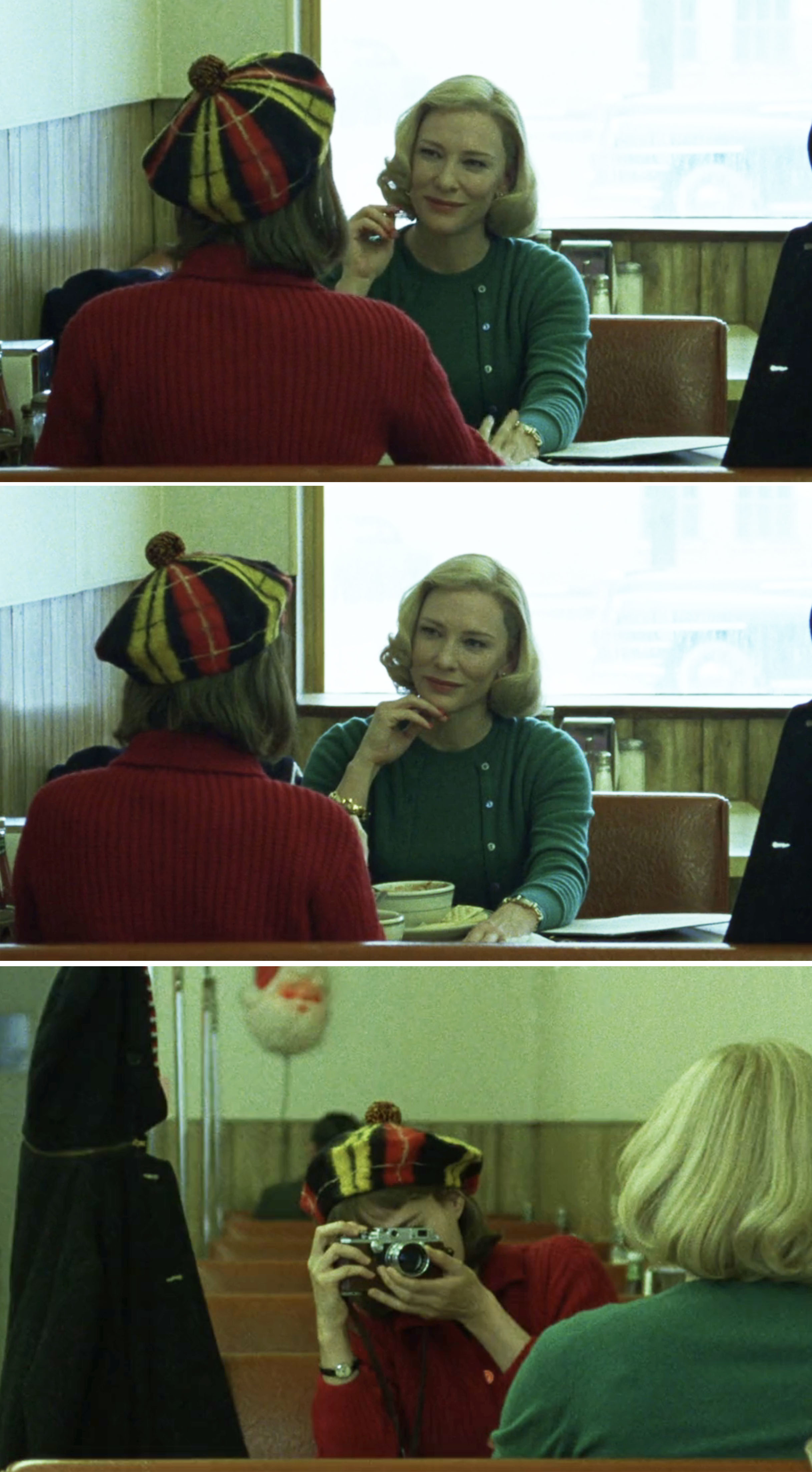 Rooney Mara and Cate Blanchett in a cafe scene from the film "Carol." Rooney wears a plaid hat, Cate wears a green cardigan. Rooney takes a photo of Cate