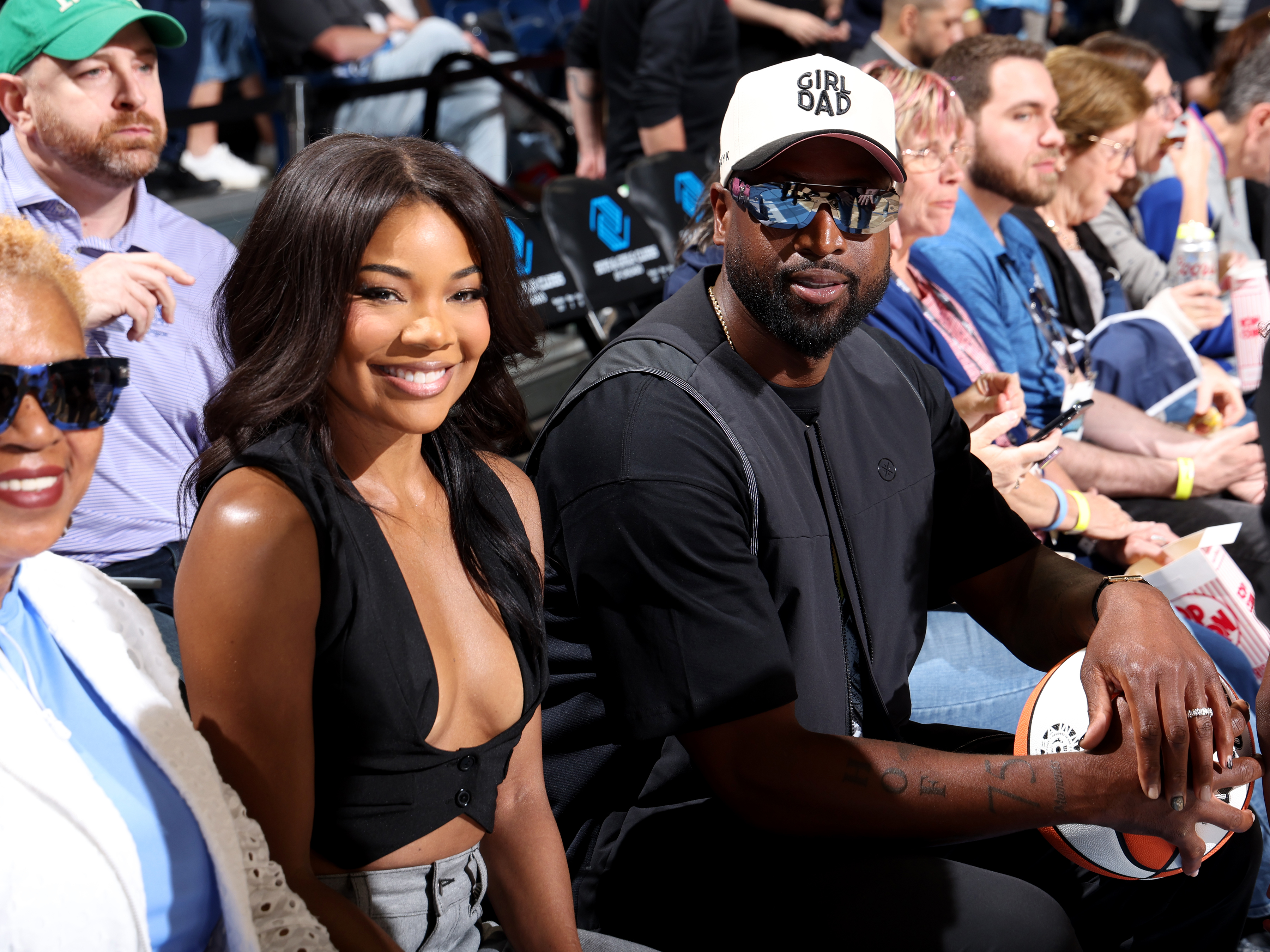 Gabrielle Union and Dwyane Wade sit courtside at a basketball game, smiling and enjoying the event. Dwyane wears a "Girl Dad" hat and sunglasses