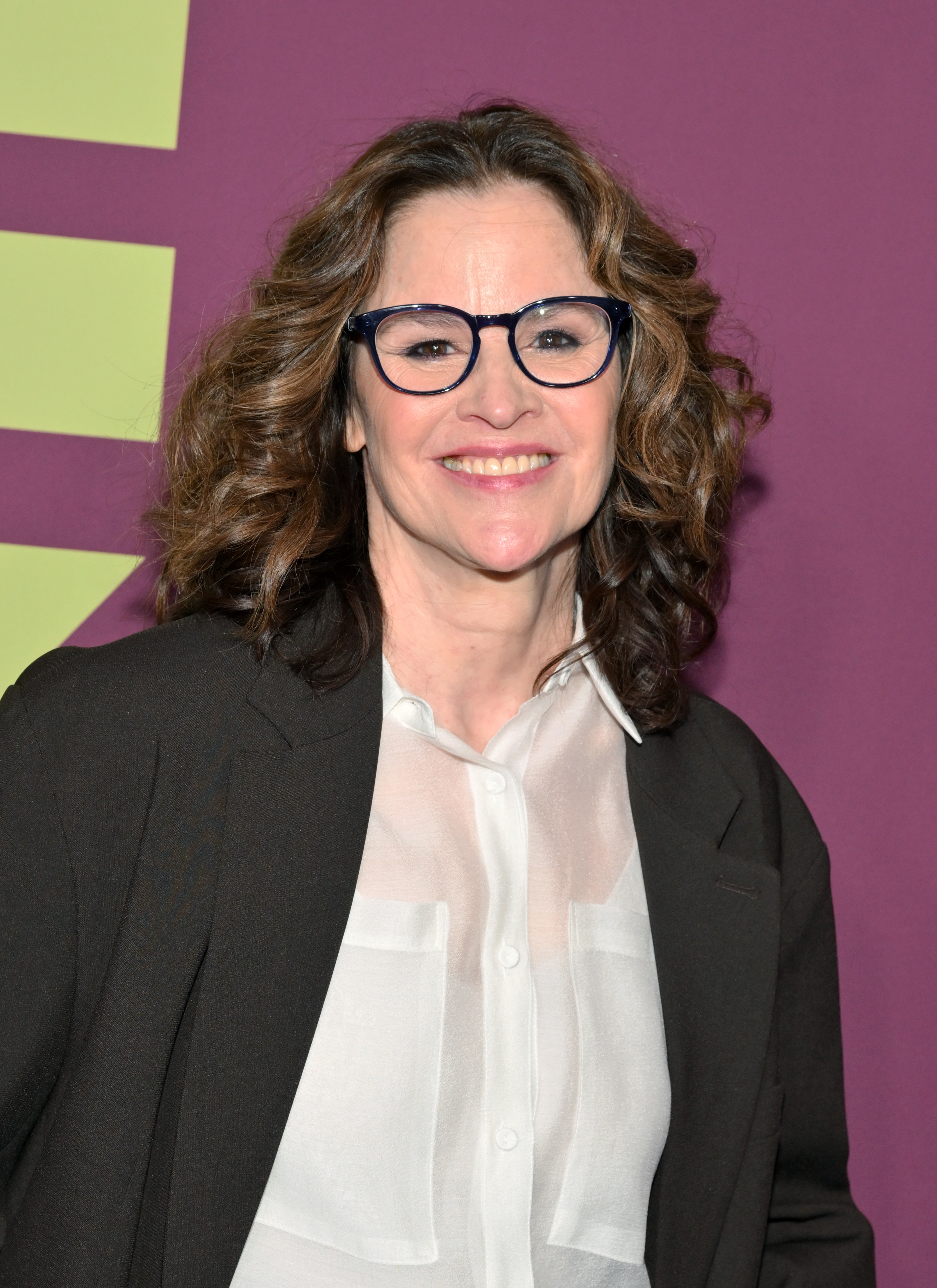 A woman with curly hair, wearing glasses, a white shirt, and a dark blazer smiles in front of a backdrop