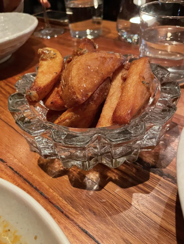 A dish of thick-cut potato wedges served in a decorative glass bowl on a wooden table, with various glasses and dishes in the background