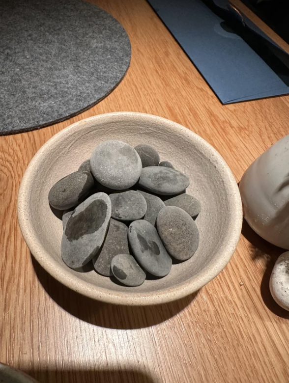 A bowl filled with smooth, gray stones is placed on a wooden table. The table also has a stone decoration and part of a circular fabric mat