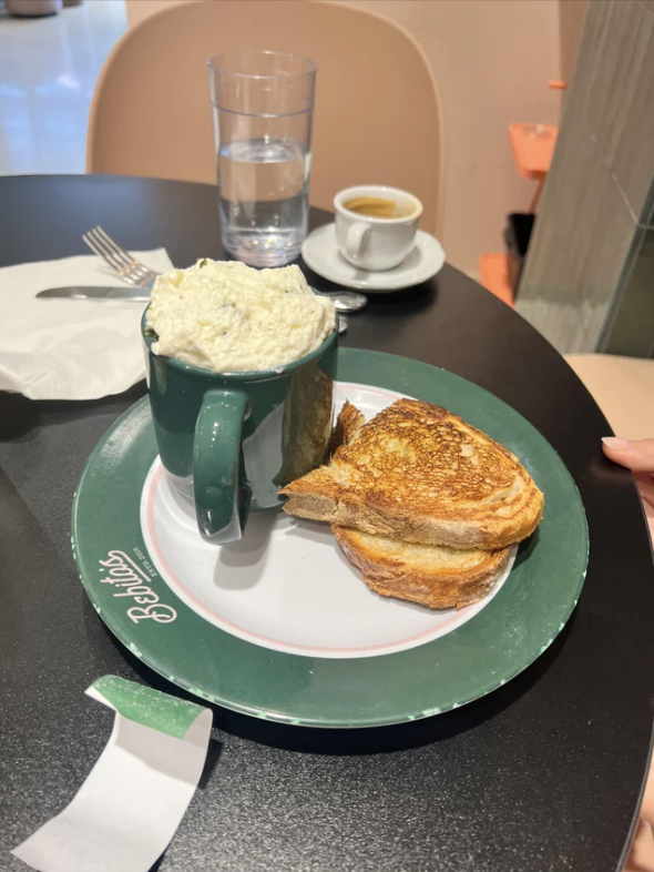 A plate with grilled cheese sandwiches and a green mug overfilled with soup at a table in a cafe. A glass of water and espresso cup are in the background