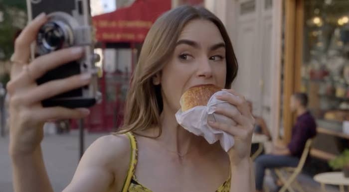 Lily Collins takes a selfie while biting into a pastry on a street with outdoor seating in the background