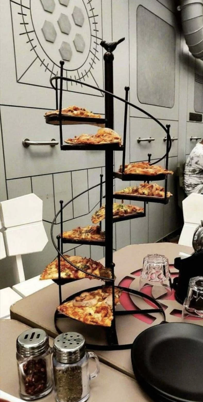 A spiral staircase pizza holder with slices of various pizzas on each level, placed on a table set for dining with plates, glasses, and condiments