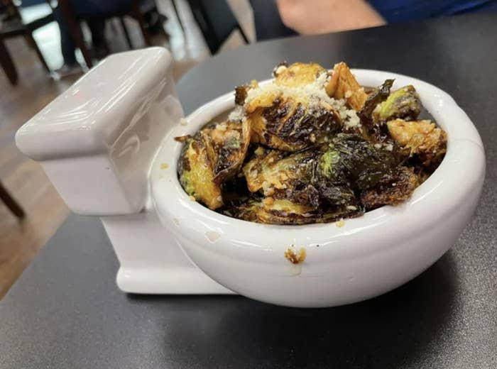 A quirky bowl shaped like a toilet filled with roasted Brussels sprouts, garnished with grated cheese, on a dining table