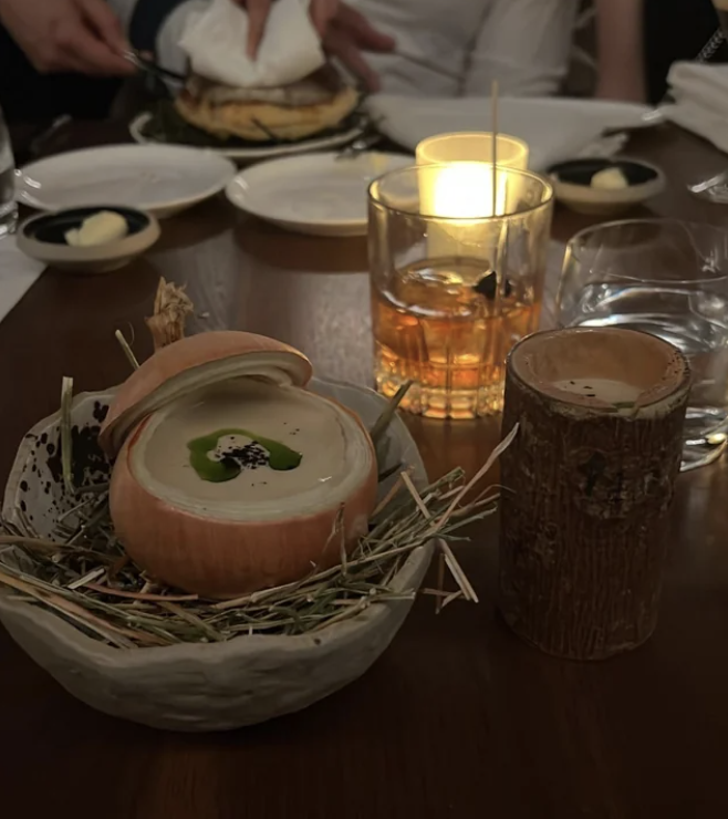 An artistic dish is served in a unique bowl, with a drink in a rustic cup next to it, on a restaurant table setting. Diners are visible in the background