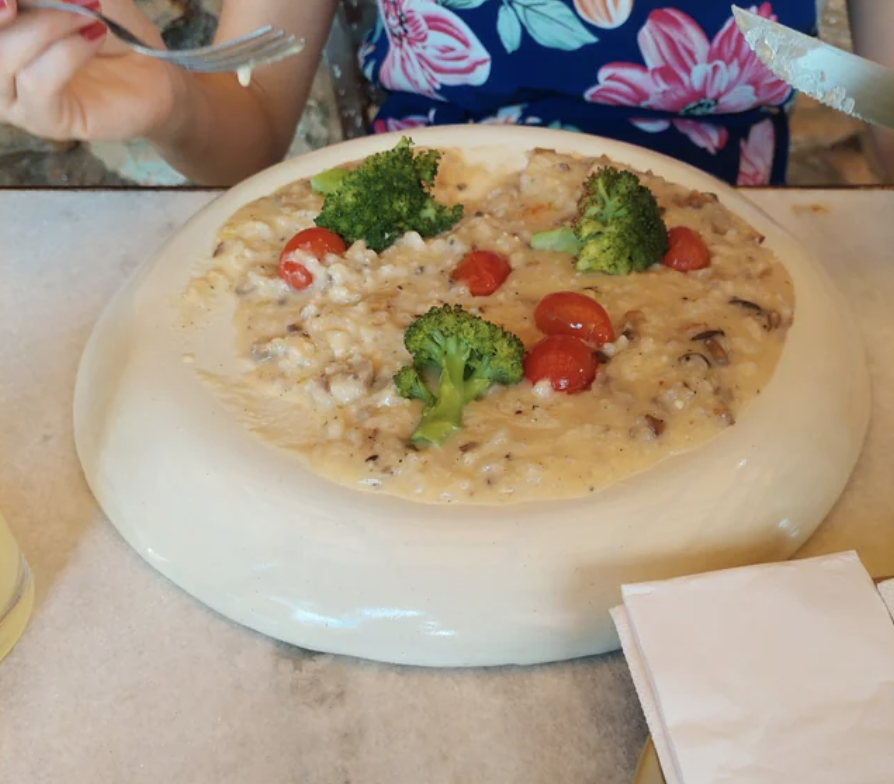 A person is about to eat a dish of risotto topped with broccoli and cherry tomatoes, placed on a white plate. They are holding a spoon