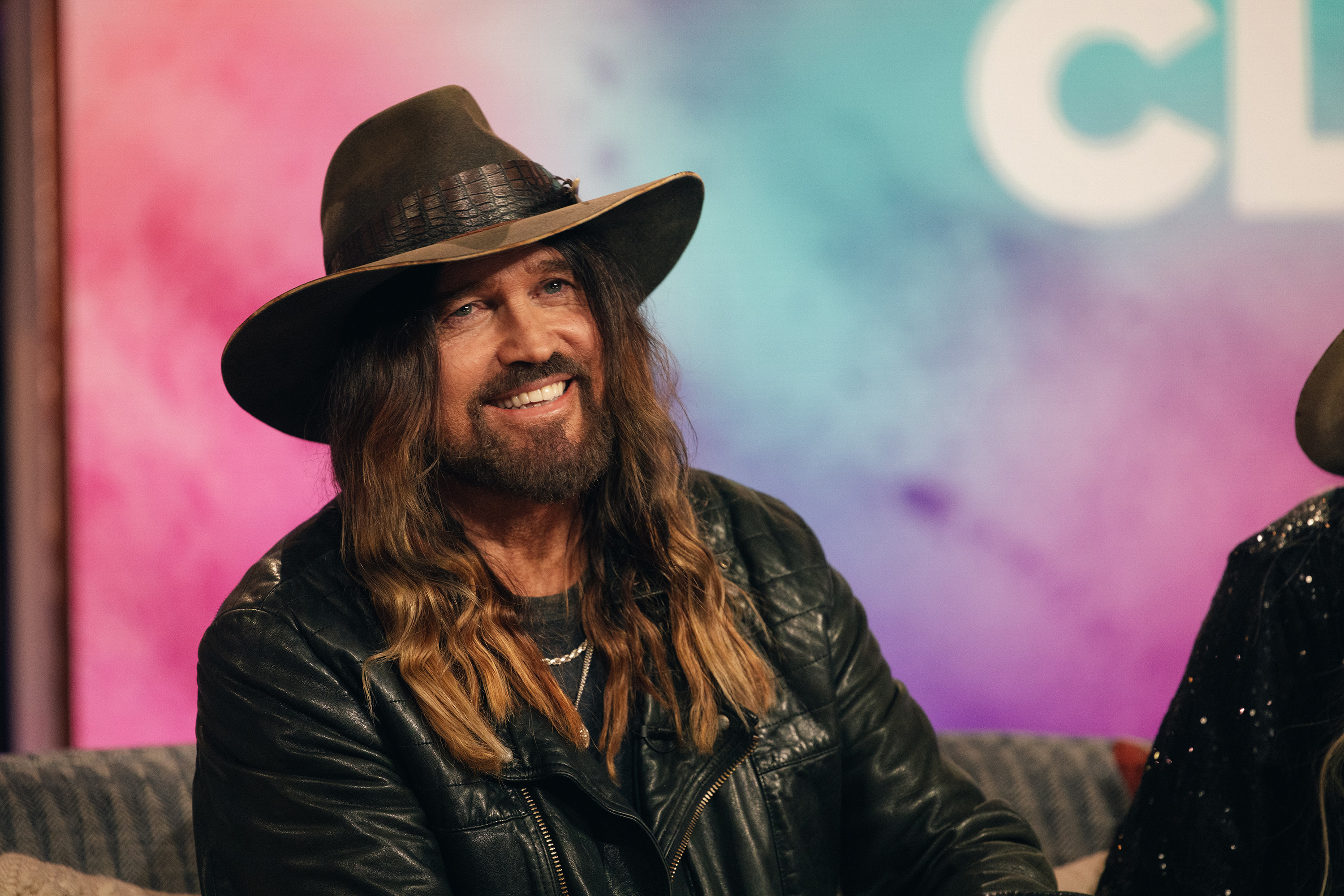 Billy Ray Cyrus sits, wearing a leather jacket and wide-brimmed hat, smiling on a talk show set with a colorful backdrop