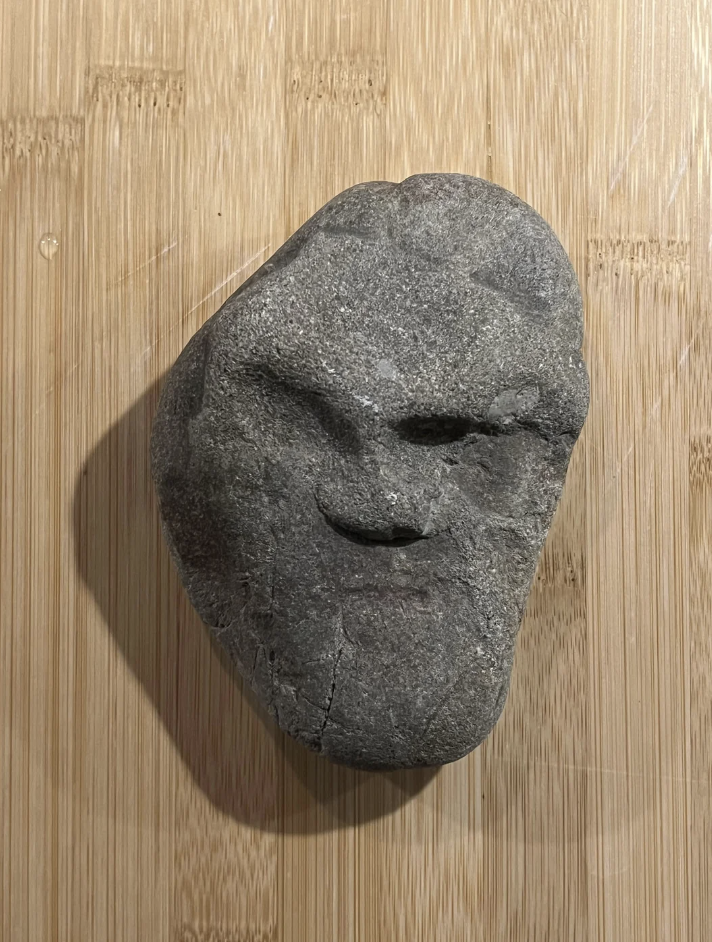 A rock resembling a human face with defined eyes, nose, and mouth sits on a wooden surface