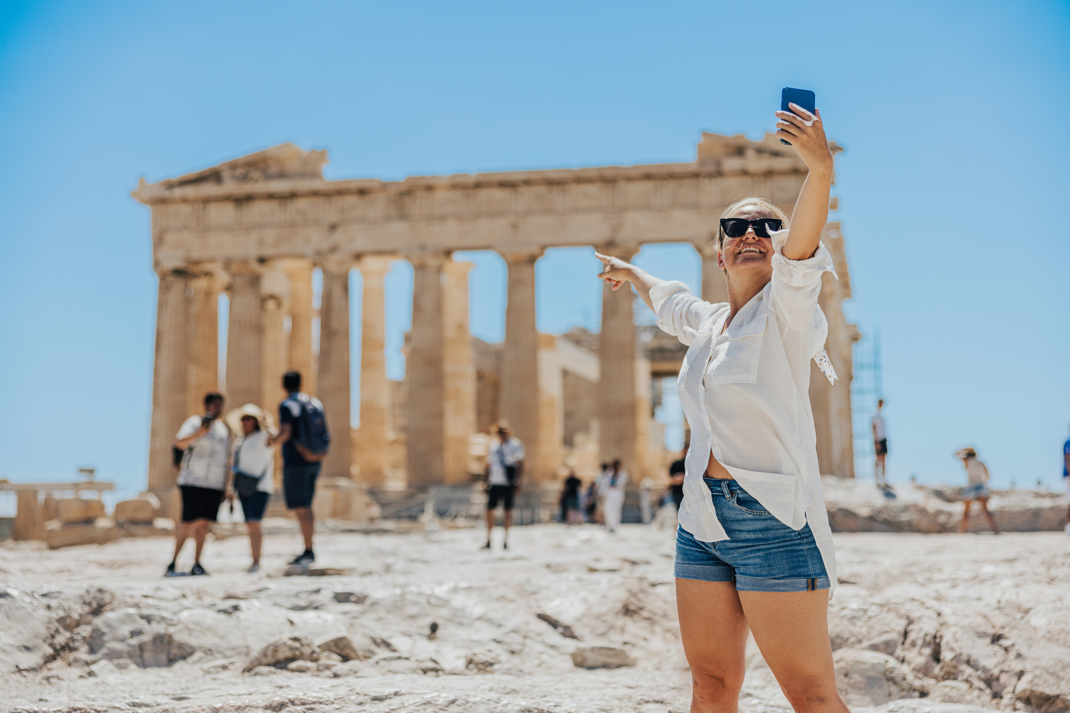 A woman wearing sunglasses, a white shirt, and denim shorts takes a selfie while pointing at ancient ruins with columns in the background. Tourists are nearby