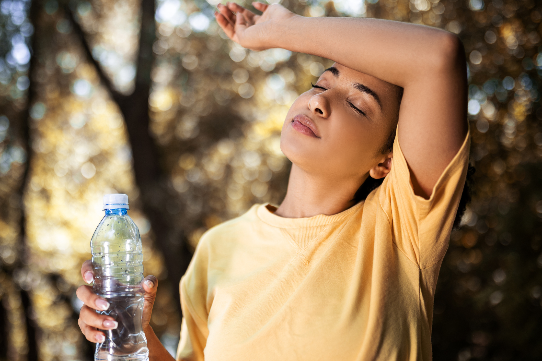 A woman in a yellow shirt appears tired and holds a water bottle, wiping her forehead with her arm, likely after exercising or being in the heat