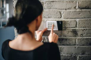 A person is seen from behind typing on a wall-mounted keypad, possibly for a home security system. The brick wall is visible. The person's face is not shown