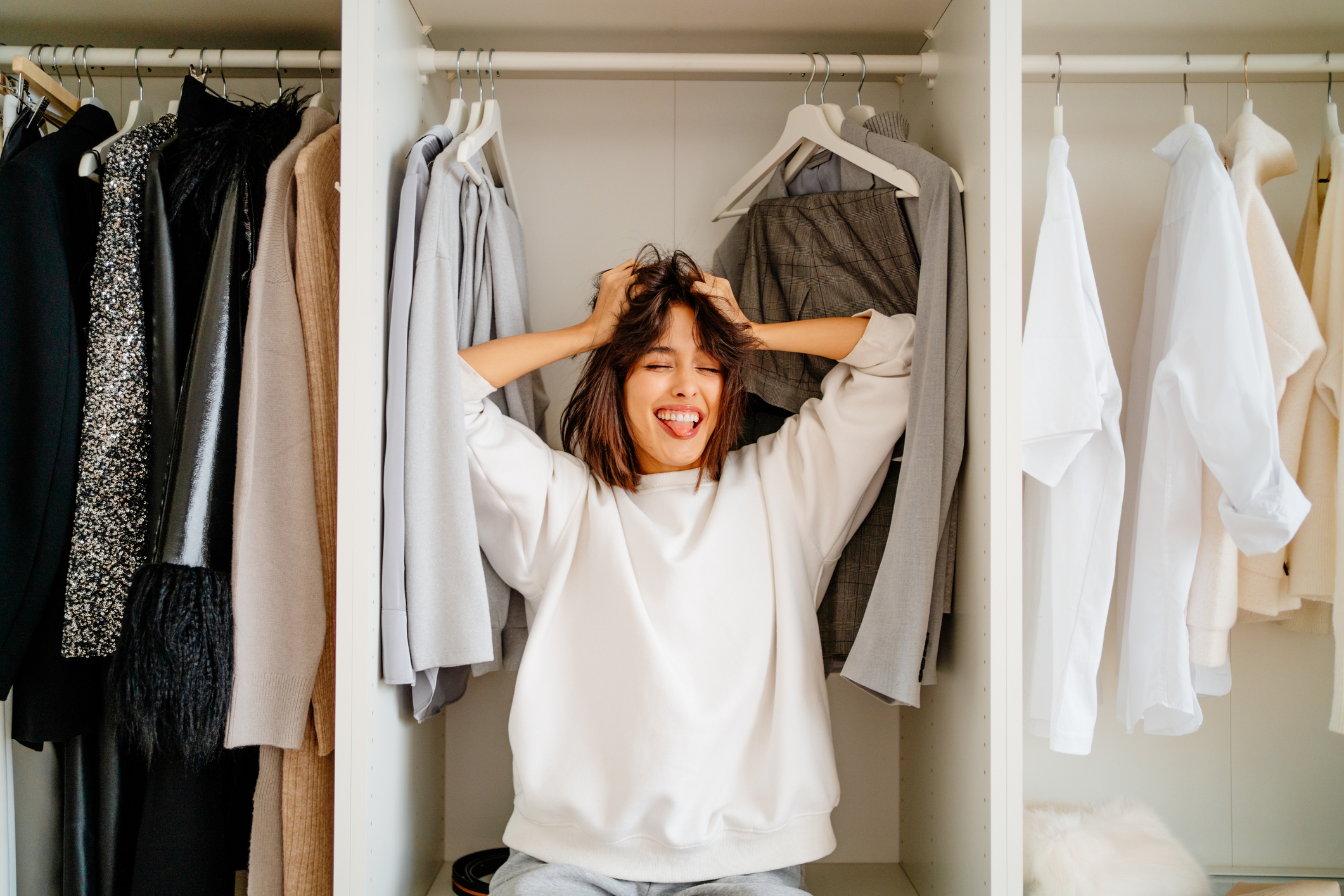 A person sits inside a wardrobe, holding their head and sticking out their tongue, surrounded by various clothes on hangers