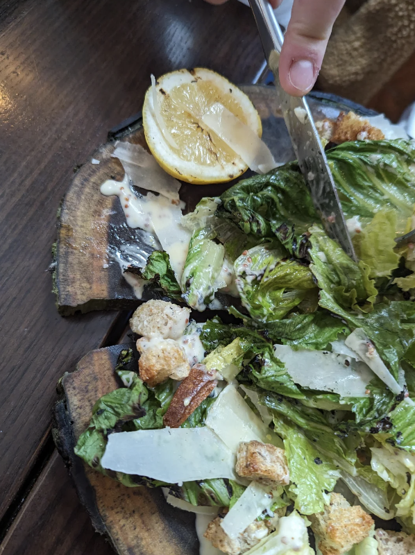 A close-up of a half-eaten Caesar salad with a lemon wedge, croutons, lettuce, and shaved cheese on a dark wooden table. A hand is seen holding a knife