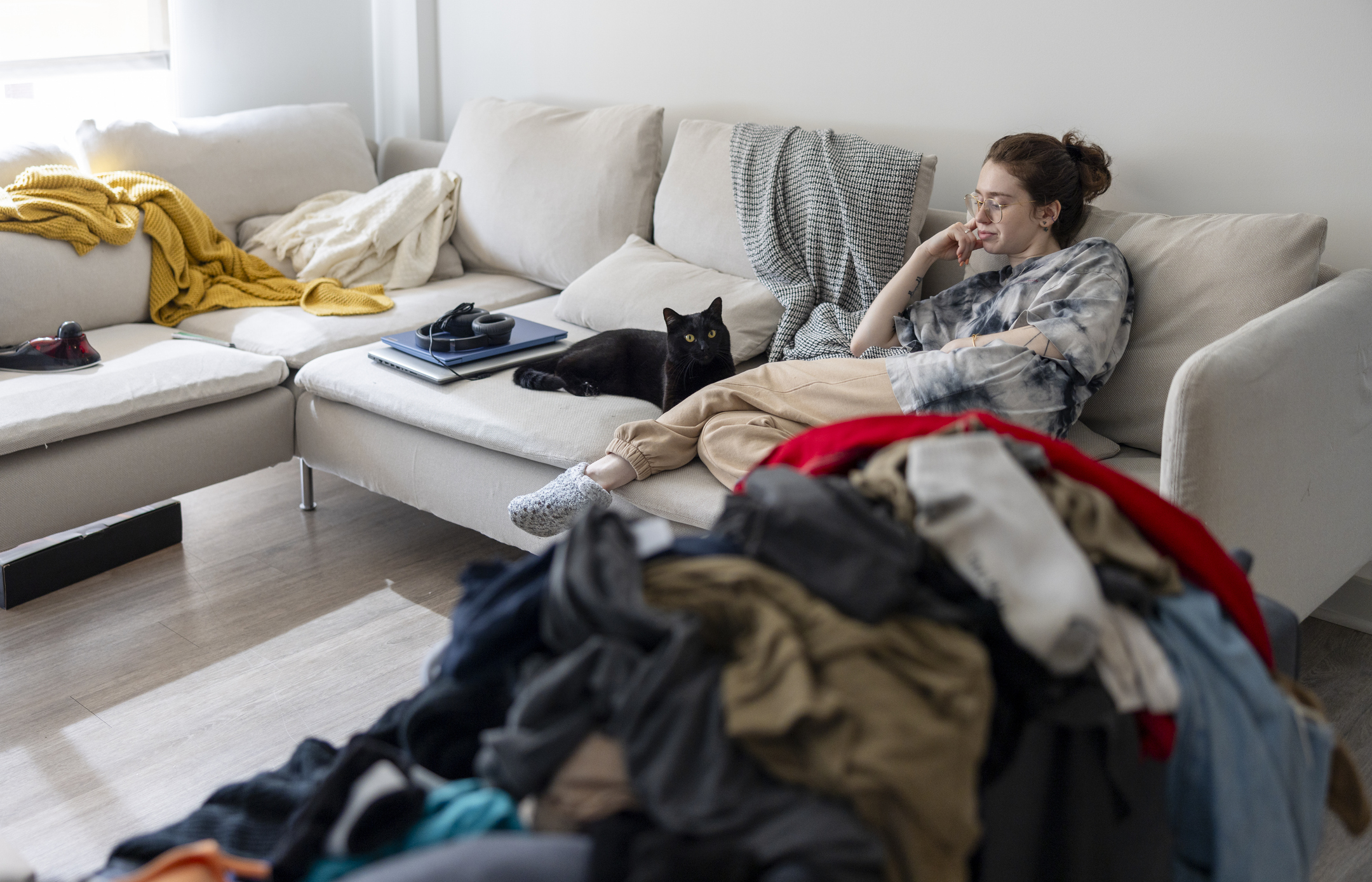 A woman sits on a couch beside a black cat, surrounded by a messy pile of clothes in a living room