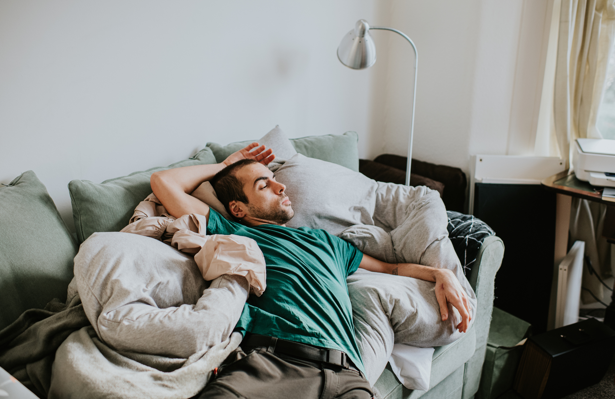 A man in a green shirt is lounging on a couch surrounded by pillows and blankets, appearing relaxed