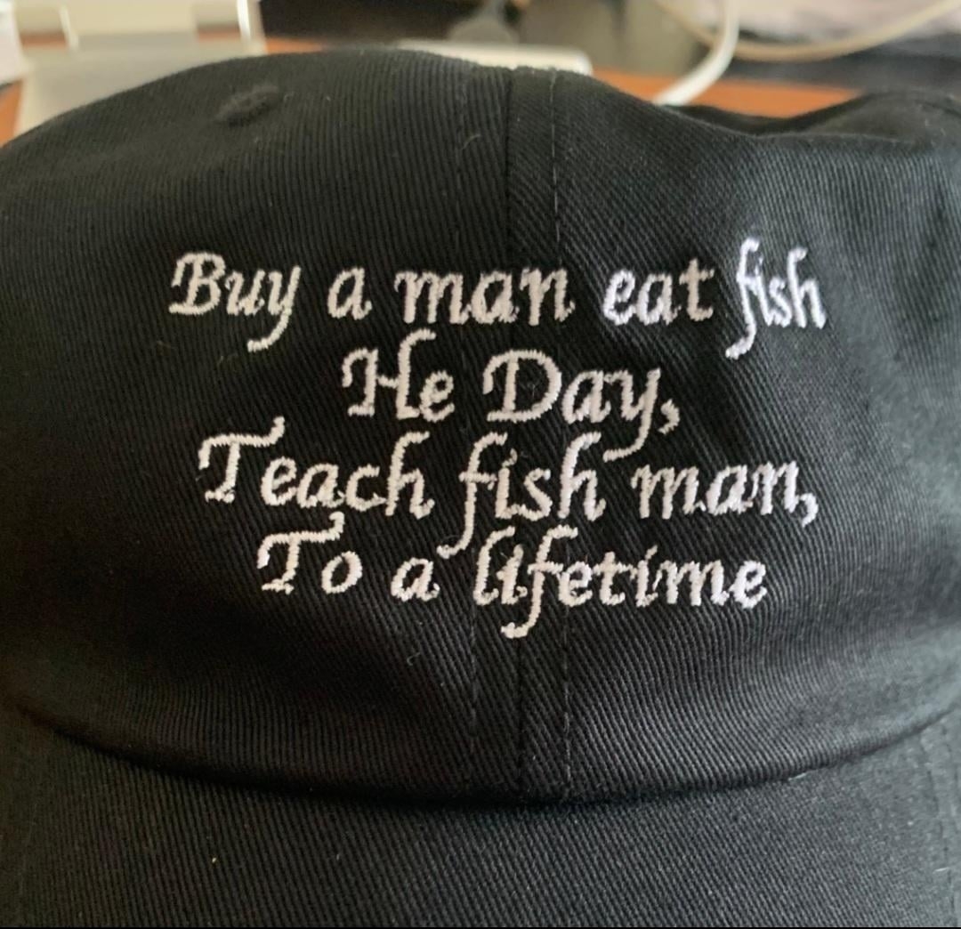 Baseball cap with embroidered text: "Buy a man eat fish He Day, Teach fish man, To a lifetime."