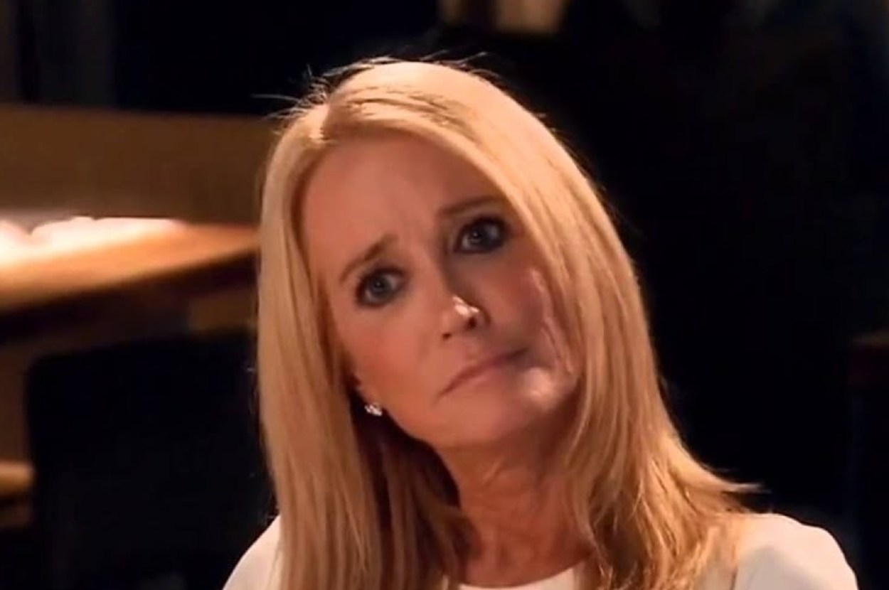 Kim Richards in a white top, seated at a restaurant table, looking intently at someone. Visible #RHOBH tag and Bravo network logo on the screen