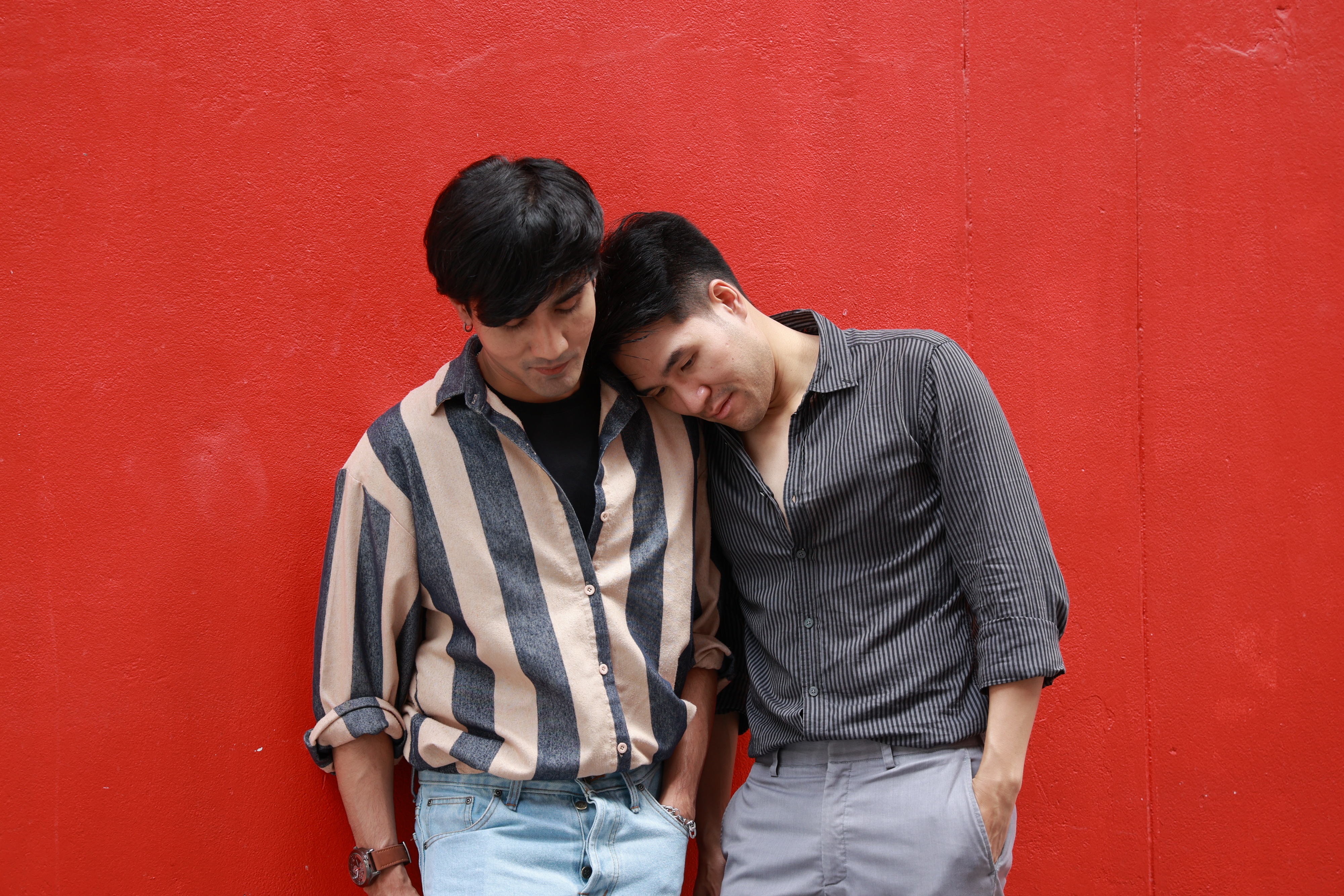 Two individuals stand against a red wall, wearing casual button-down shirts. One person leans their head on the other's shoulder