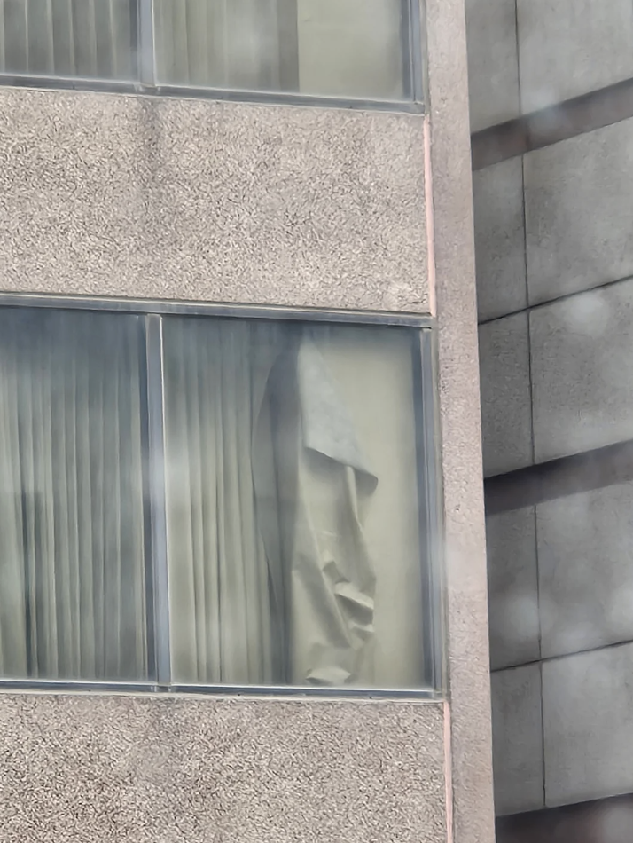 A mysterious figure appears behind a partially open curtain in a building's window, creating a curious and eerie visual effect