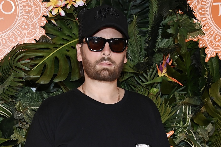 Scott Disick wearing a black t-shirt and sunglasses, poses in front of a tropical-themed backdrop at the Tao Beach event