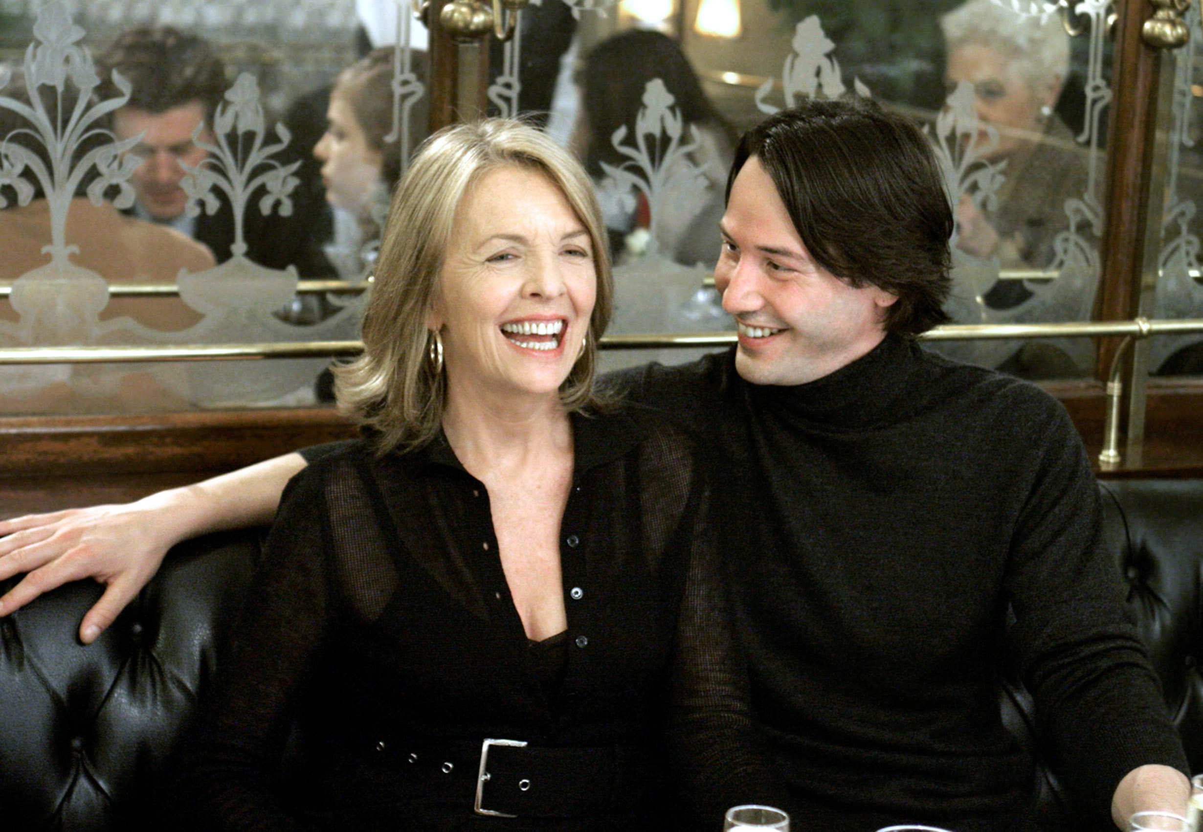 Diane Keaton and Keanu Reeves sitting closely and smiling at each other in a cozy indoor setting. Keaton wears a belted dress, and Reeves wears a turtleneck