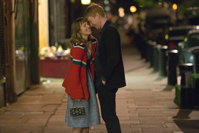 Rachel McAdams and Domhnall Gleeson share a tender moment on a dimly lit street. Rachel is wearing a cozy coat over a dress and holding a small handbag and Domhnall is wearing dark clothing