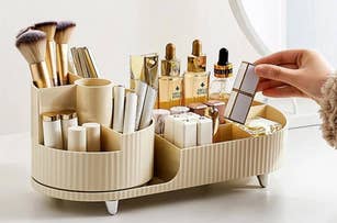 Makeup organizer holding brushes, lipsticks, foundation bottles, and other beauty products, with a hand placing a compact in the tray