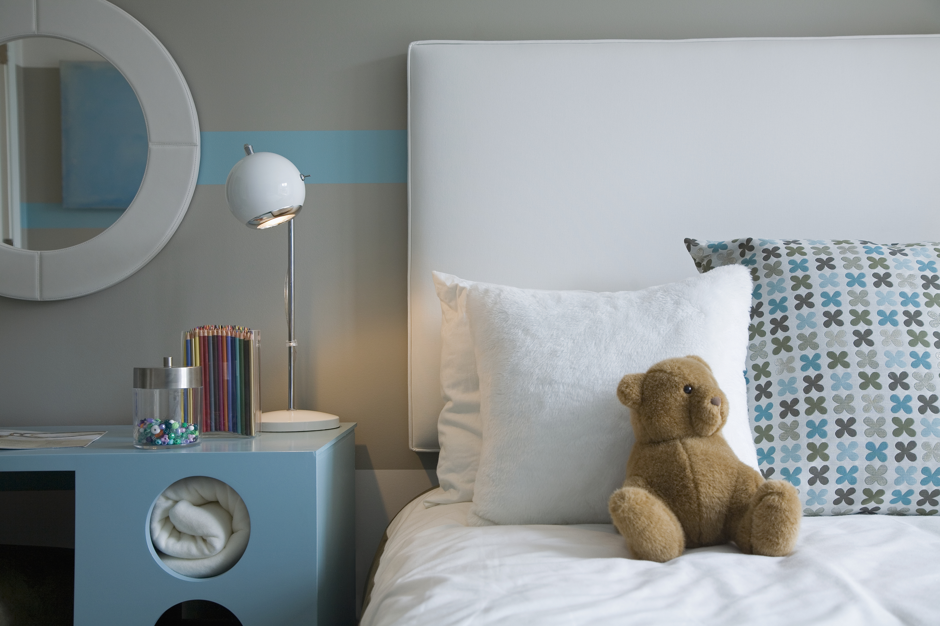 A neatly made bed with white bedding, a white and patterned pillow, and a brown teddy bear. A nightstand holds a lamp, colored pencils, and a candy jar