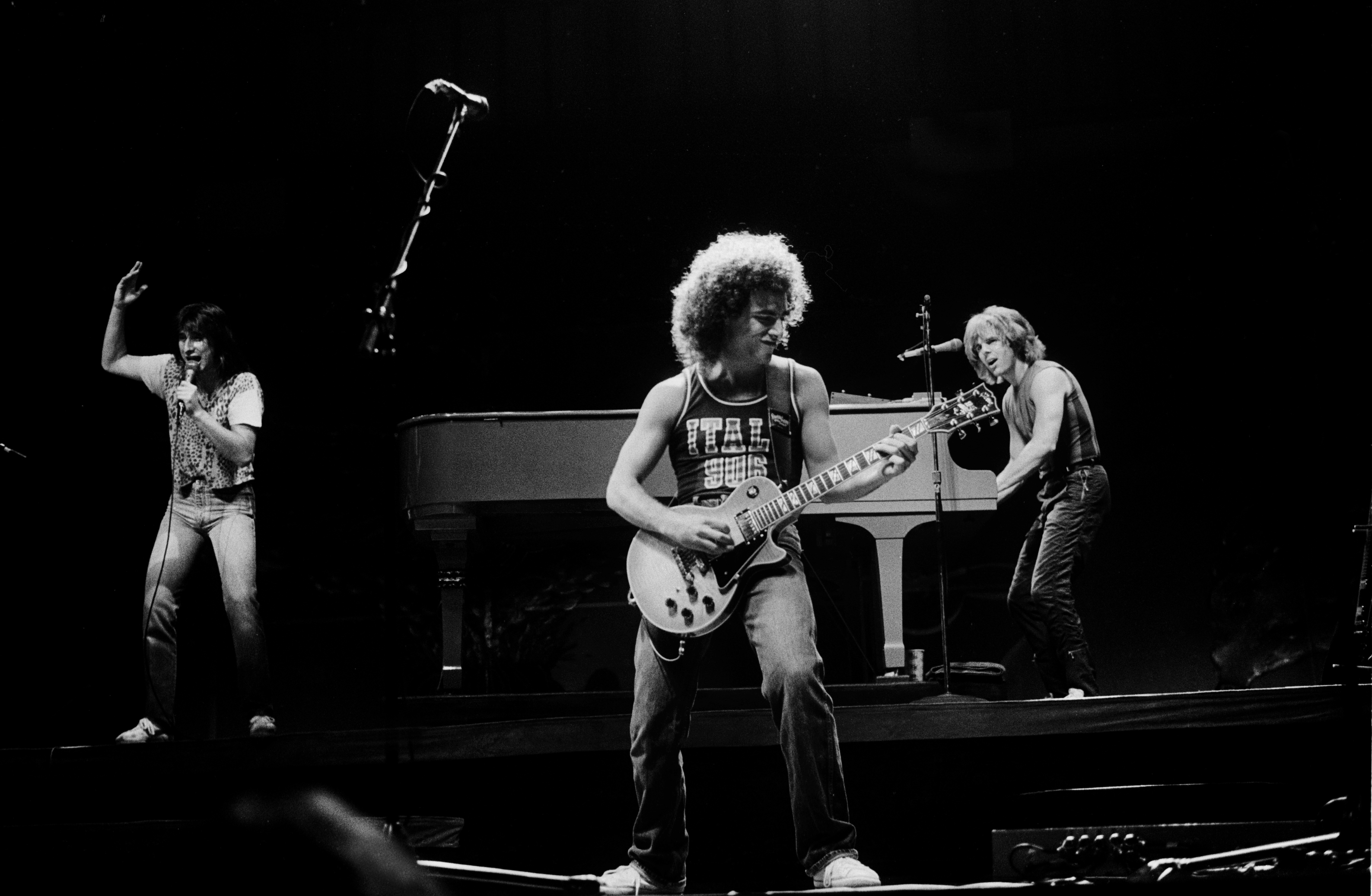 Three musicians perform on stage, one on electric guitar centrally, another dancing on the left, and the third playing keyboards on the right