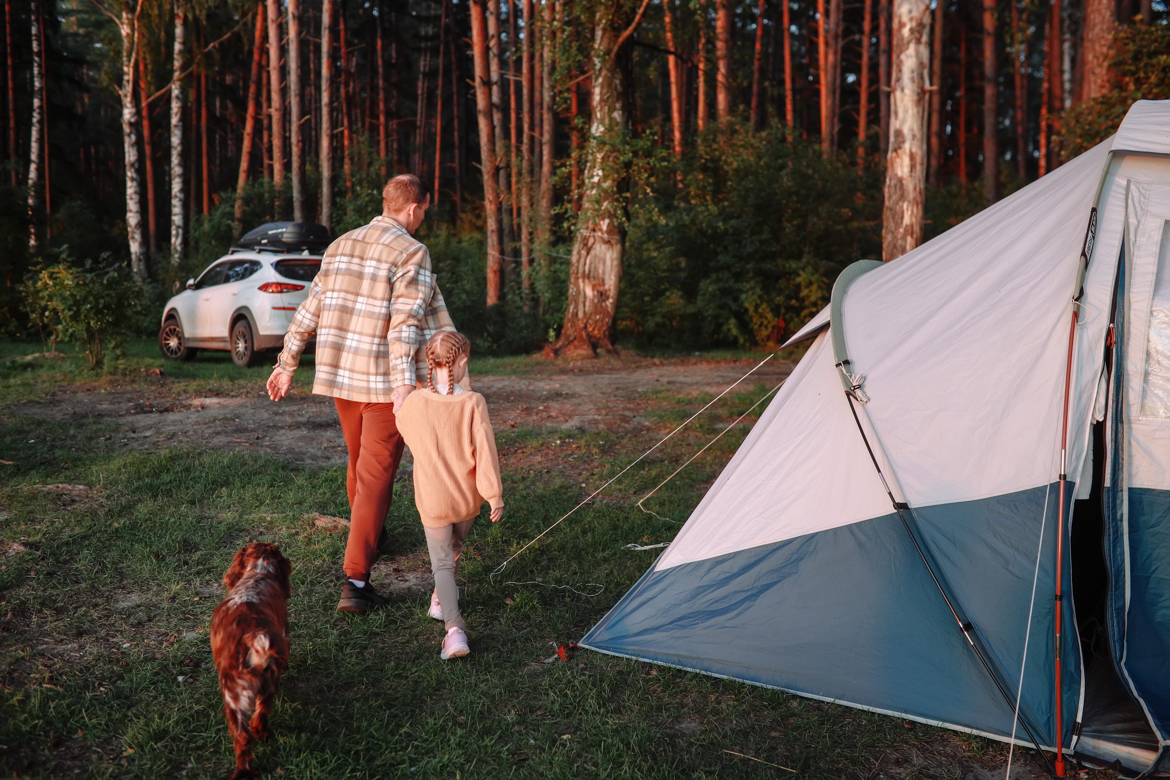 A person in a plaid shirt and child walk hand-in-hand towards a tent in a wooded campsite, with a dog following them. A car is parked in the background