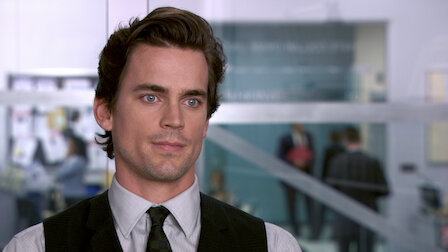 Matt Bomer wearing a suit and tie, seen from the shoulders up, with an office background where people appear to be working and conversing