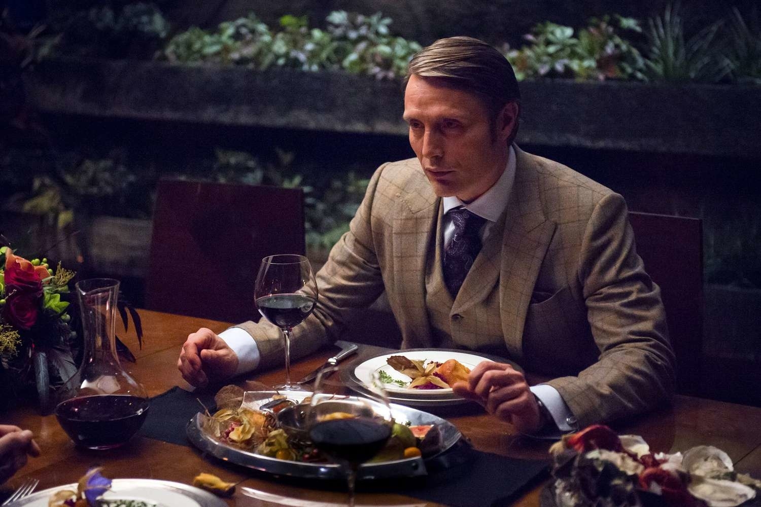 Mads Mikkelsen in a formal suit, seated at a dining table with a glass of wine and gourmet dishes in front of him