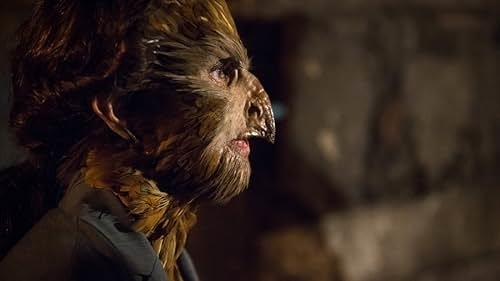 A person in detailed prosthetic makeup resembling a bird-like creature, with feathers and a beak, looking to the side in a dimly lit setting