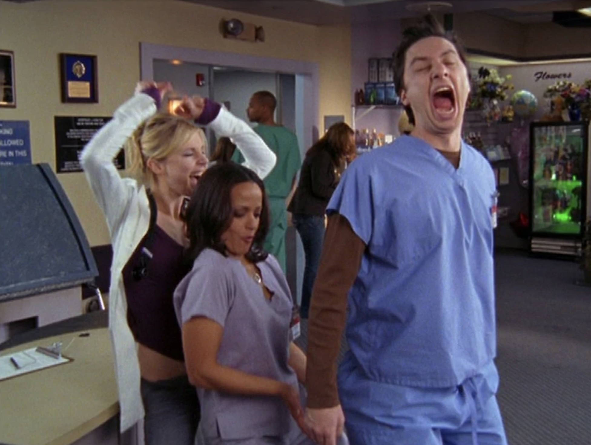 Sarah Chalke and Judy Reyes, in scrubs, humorously grab Zach Braff, who is also in scrubs, in a playful scene in a hospital setting with people in the background