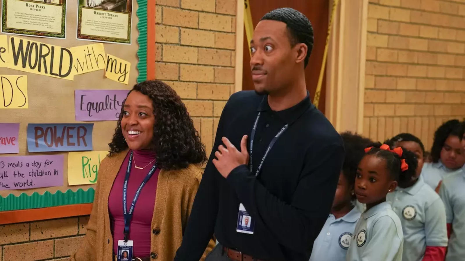 Quinta Brunson and Tyler James Williams stand in a school hallway with children in uniforms, smiling and looking at a display board