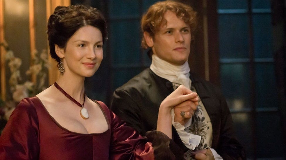 Caitríona Balfe in a period dress holding hands with Sam Heughan, who is dressed in historical attire, standing in a lavishly furnished room